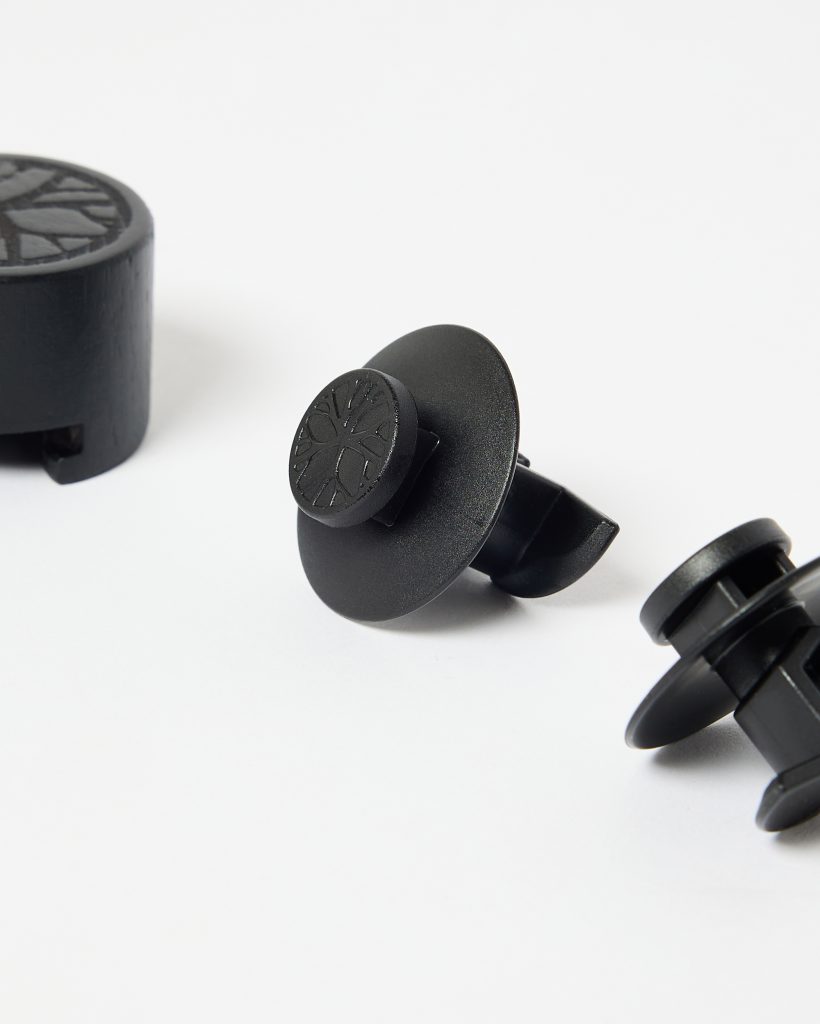 Close-up of WoodUpp accessories: two black plastic vent mounts with wide round flanges and split prongs, each topped by a small circular cap embossed with WoodUpp’s tree logo, alongside a separate black wooden puck-style cap also carved with the same tree emblem.