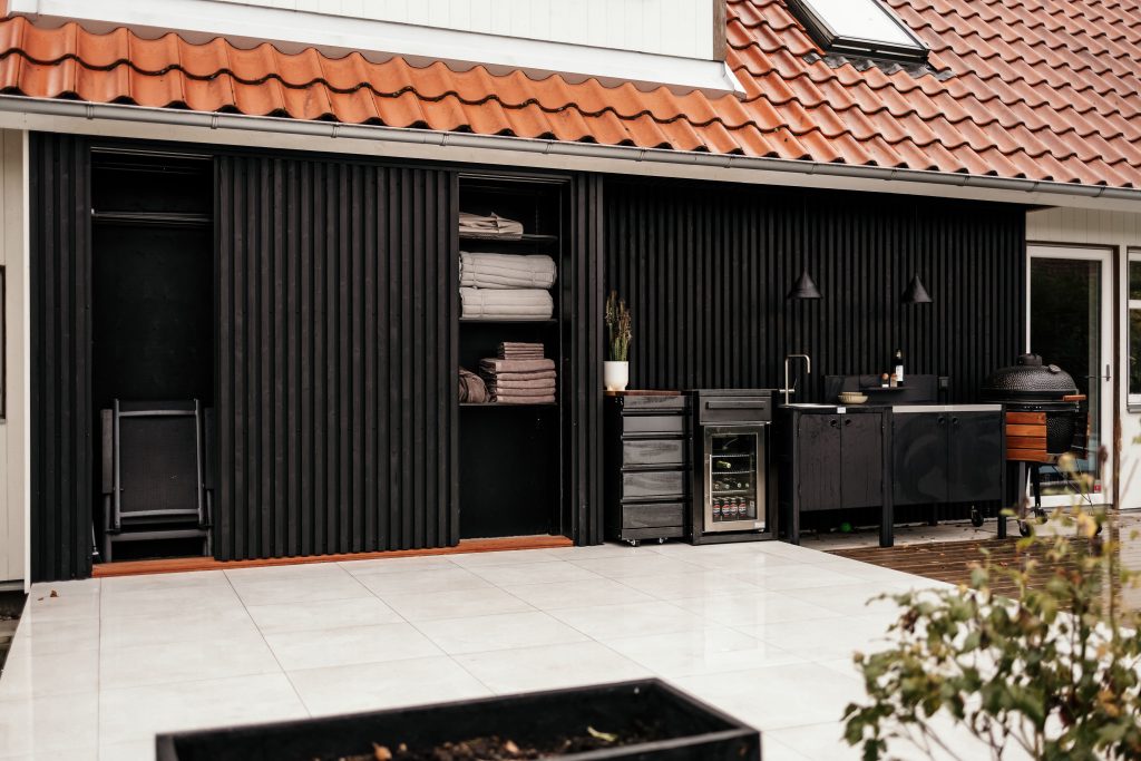 WoodUpp outdoor kitchen setup with black vertical slatted WoodUpp cladding, an integrated WoodUpp cabinet system featuring stacked drawers on casters, a compact undercounter beverage fridge, open WoodUpp shelving holding folded towels, a sink-equipped countertop, and a charcoal kamado-style grill beside the unit.