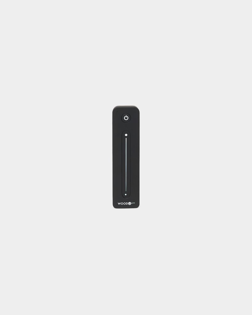 Centered on a pale background, a WoodUpp black remote control is shown: a slim rectangular unit with a circular power button at the top, a vertical touch-sensitive LED slider in the middle, two small indicator LEDs, and the WoodUpp logo printed at the bottom.