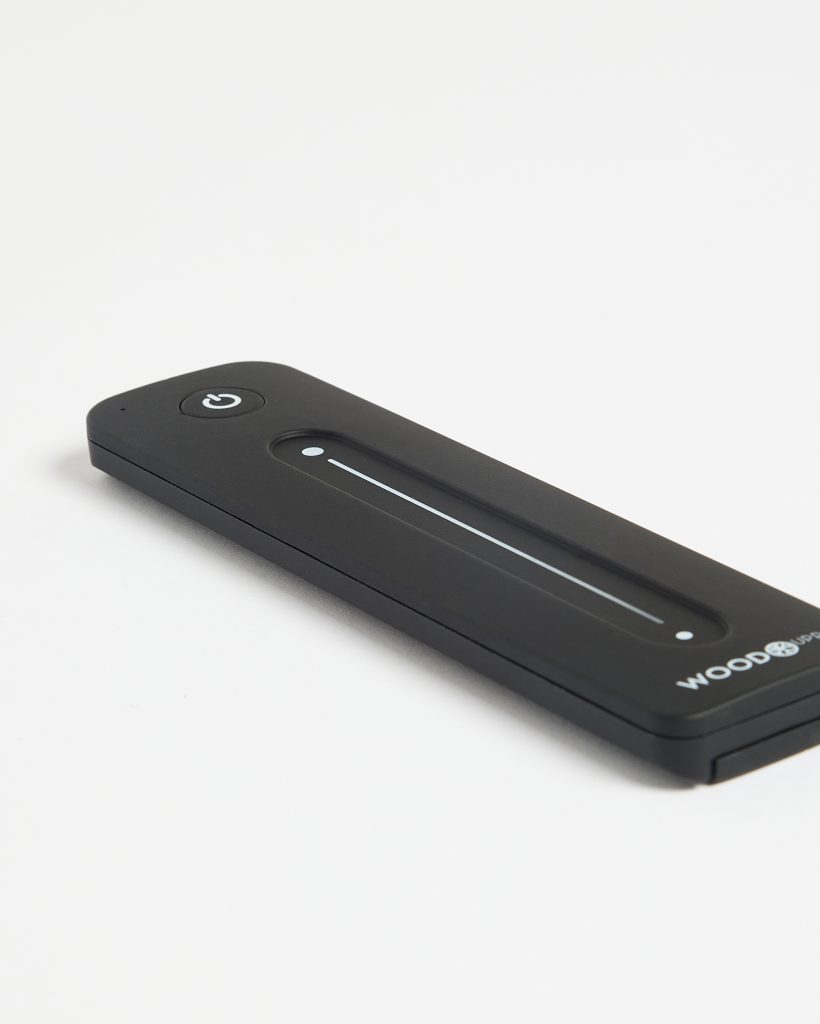 Close-up of a WoodUpp-branded slim black rechargeable electric lighter laid on a white background. The rectangular device features a circular power button, recessed touch slider with a thin illuminated line and two indicator dots, and the WOODUPP logo printed on the lower corner.