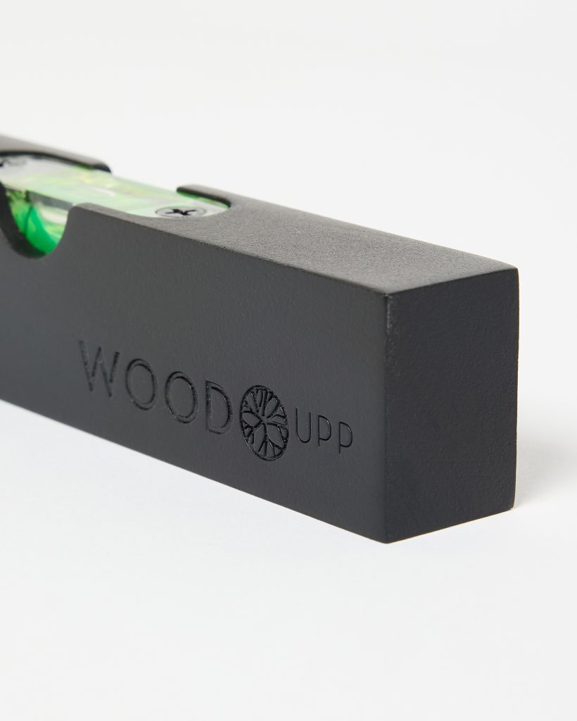 Close-up of a matte black WoodUpp spirit level: a rectangular wooden block with the WOODUPP logo and circular wheel emblem engraved on the side, featuring an inset green bubble vial secured by visible screws in a recessed channel along the top edge.