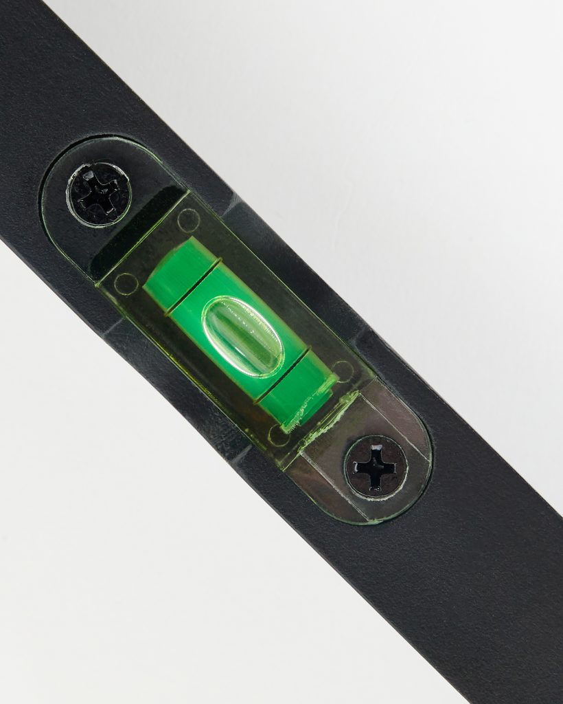 Close-up of a WoodUpp spirit level: a compact black metal bar with an integrated green bubble vial in translucent housing, the central bubble visible and secured by two Phillips-head screws. Matte-finished, diagonally oriented against a clean white background.