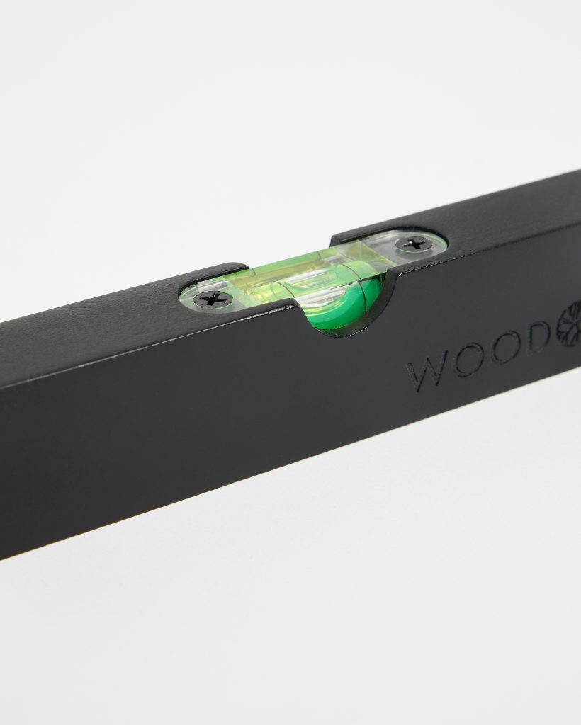 Close-up of a WoodUpp wooden spirit level: a matte black rectangular body with an inset transparent green bubble vial, visible air bubble centered between lines, secured by two metal screws, and the WoodUpp logo subtly embossed on the side finish.