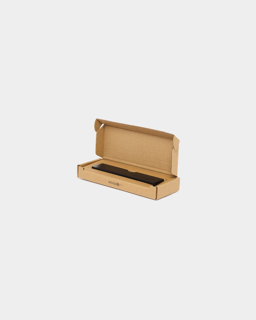 Open recycled cardboard packaging branded WoodUpp displays a slim black rectangular product — a matte-finish bar resting in a fitted tray. The kraft box lid is open, showing the WoodUpp logo on the box front and the black bar accessory nestled securely inside.