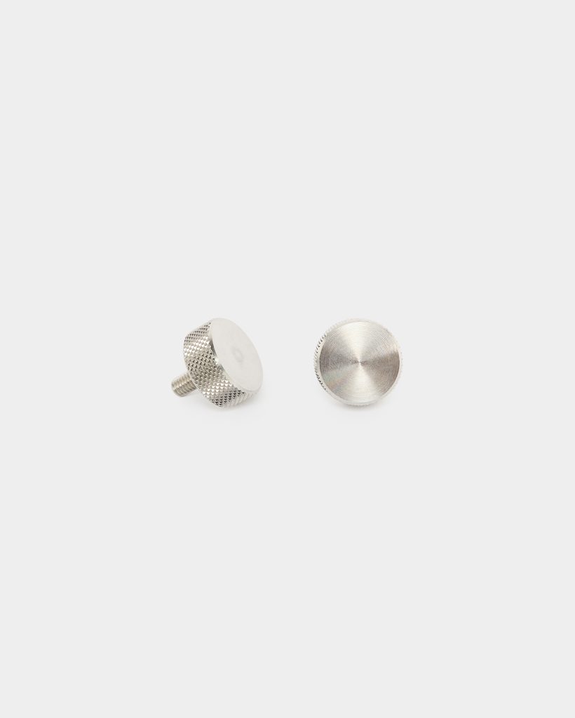 WoodUpp: two small silver metal thumb screws laid on a white background. One screw lies on its side showing a short threaded stud and knurled cylindrical edge; the other displays a flat circular head with concentric machining marks. Brushed nickel finish, textured grip for hand tightening.