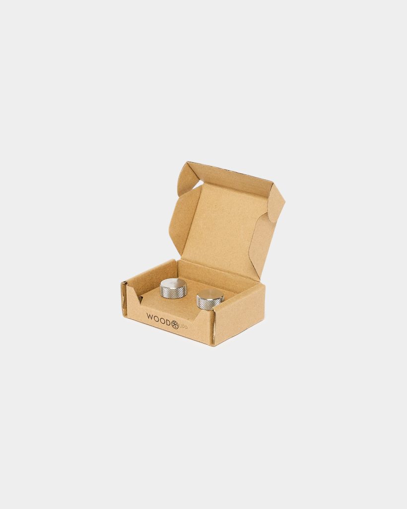 Open WoodUpp cardboard box containing two silver, knurled metal knobs placed side by side. The small brown packaging displays the WoodUpp logo on the front; the cylindrical, textured knobs have flat tops and machined side grips, photographed against a pale neutral background.