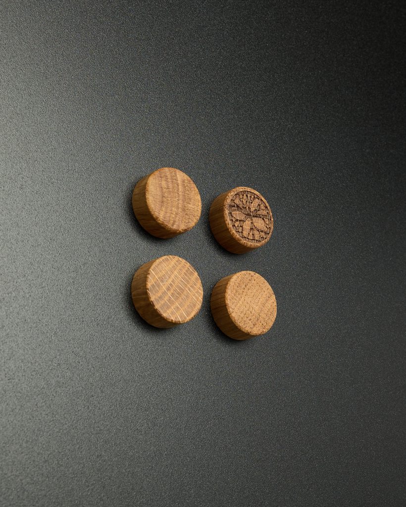 Four WoodUpp wooden magnetic discs arranged in a loose square on a dark textured background; three display smooth oak grain while the fourth is laser-engraved with the WoodUpp tree logo, warm-toned cylindrical shapes with visible growth rings and chamfered edges.