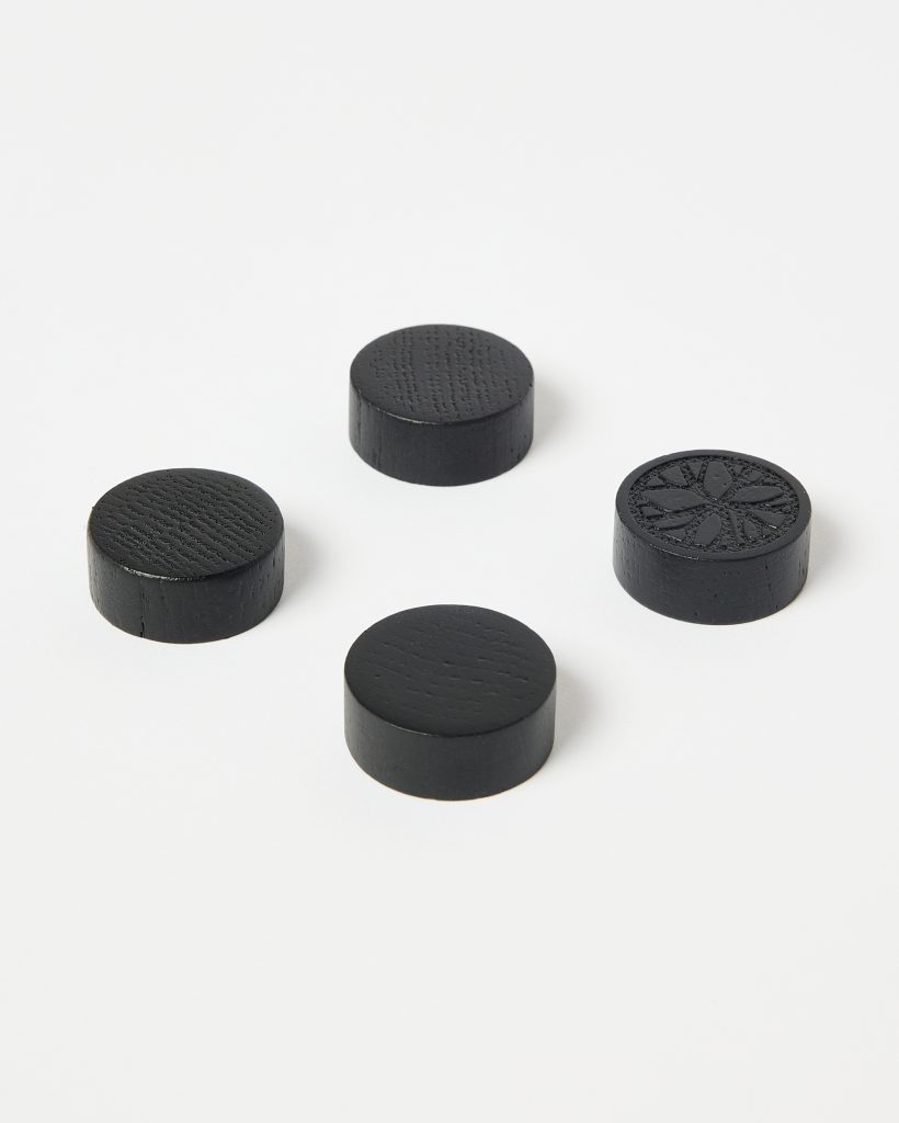 Four small round black wooden discs from WoodUpp sit on a white background: three show visible wood grain and smooth matte tops, while a fourth features a carved floral/star motif. Each cylindrical disc has flat faces and short straight sides, suggesting decorative or functional wooden plugs.