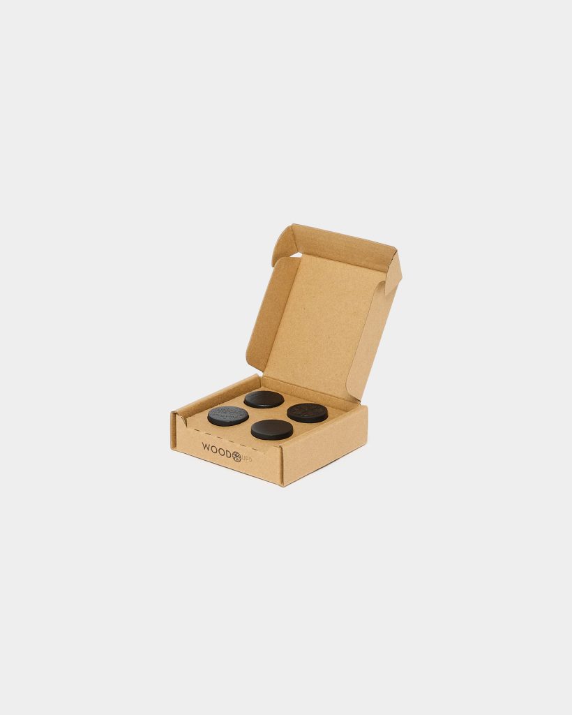 Open kraft WoodUpp box centered on white background containing four round WoodUpp floor protectors: two smooth matte black discs, one textured dark grey disc, and one patterned black disc. The compact recycled cardboard packaging is branded with the WoodUpp logo on the front.