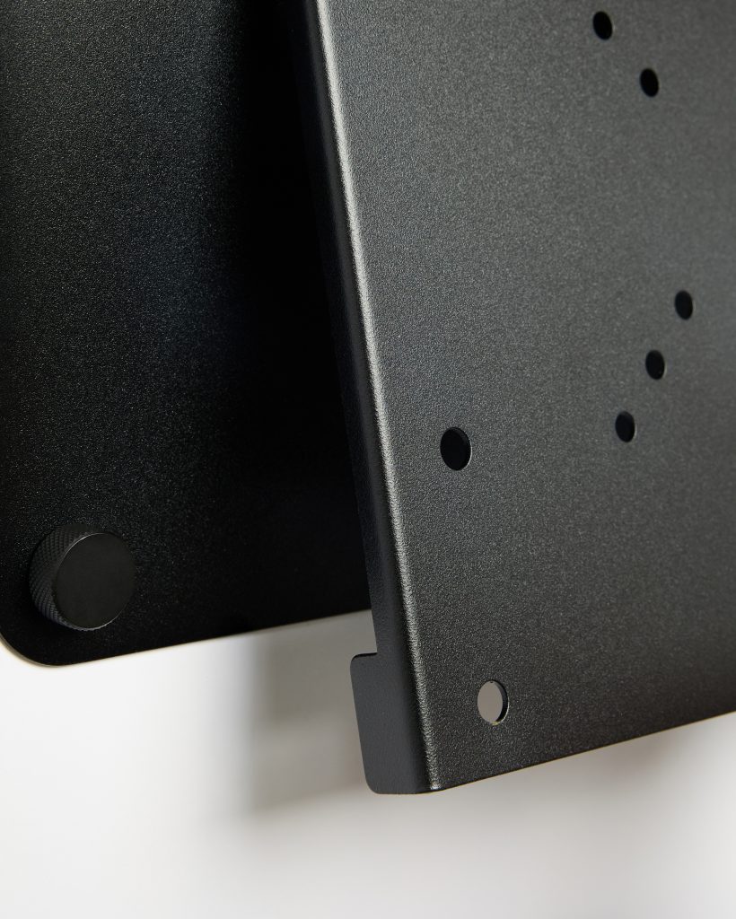 Close-up of WoodUpp black powder-coated metal monitor mounting parts: a VESA-style plate with multiple screw holes and a matching wall bracket. The image shows textured matte finish, a rounded bottom lip, and a knurled adjustment knob, highlighting solid steel construction and precise cutouts.