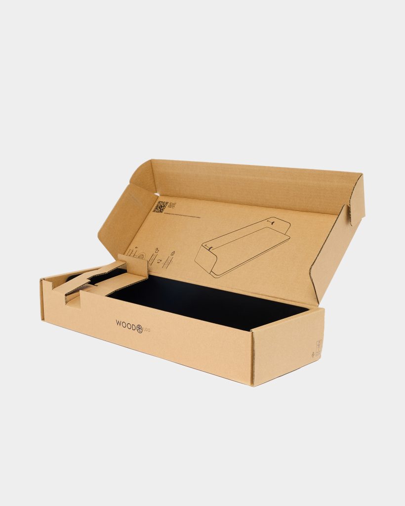 Open WoodUpp corrugated shipping box with printed WoodUpp logo, internal black protective lining, and shaped cardboard inserts. The lid shows a printed assembly diagram of a slim sloped wooden laptop stand, while the left compartment contains folded cardboard supports and small accessory cutouts for the WoodUpp product.