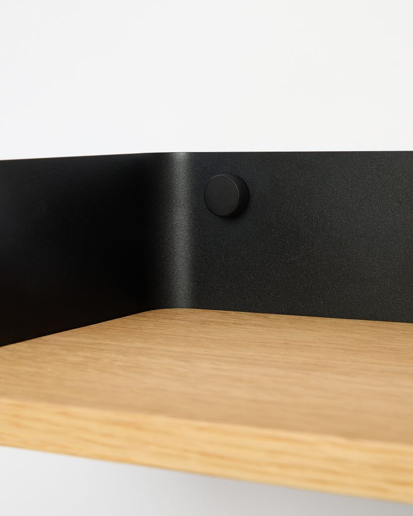 WoodUpp close-up showing a light oak wall shelf with visible grain and a clean, chamfered front edge, mounted against a textured matte-black steel backplate. The WoodUpp design includes a low-profile, knurled round black metal peg or cable grommet fixed to the plate, emphasizing functional, minimalist styling.