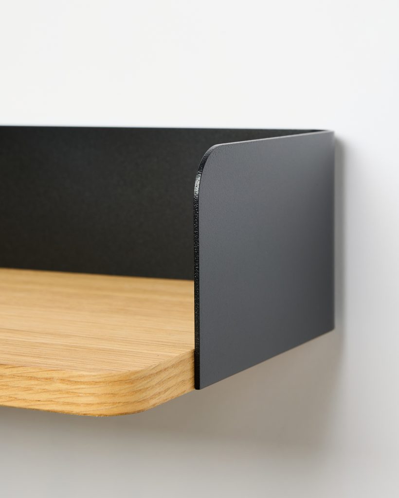 Close-up of a WoodUpp wall shelf showing a light oak shelf board with rounded front edge paired with a matte black steel side panel/rim. The WoodUpp product features a slim, curved metal bracket rising at the shelf’s rear and side, creating a minimalist floating-shelf detail.