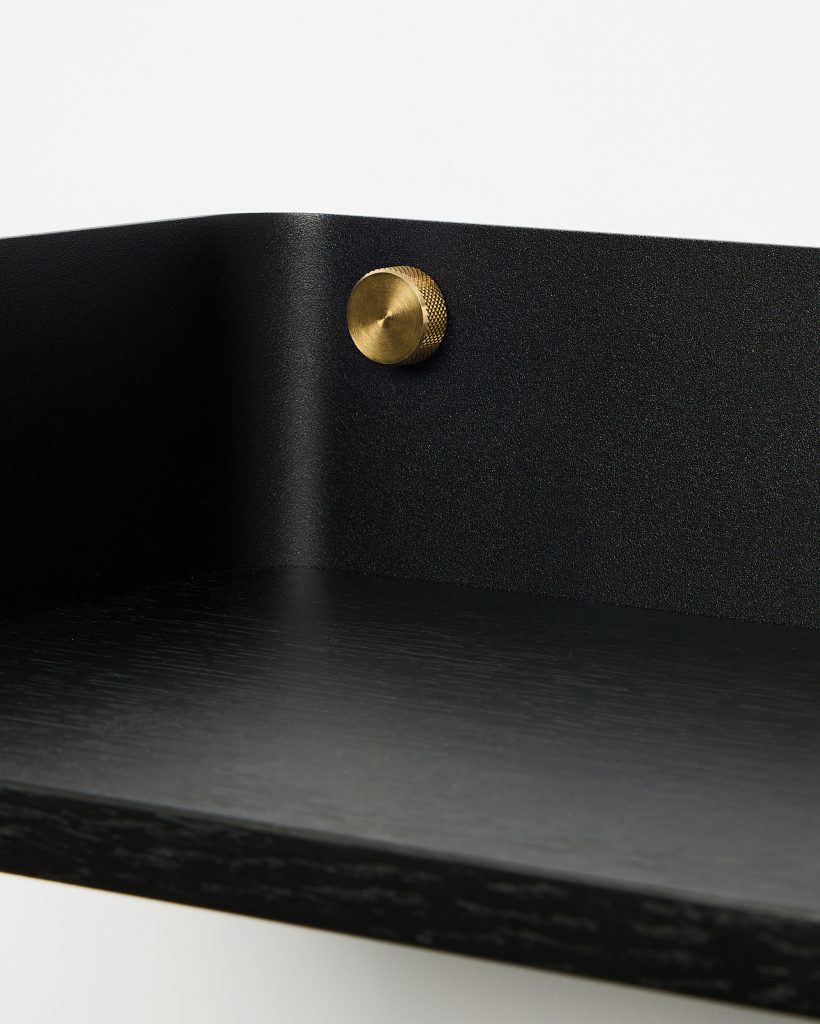 Close-up of a WoodUpp wall shelf showing a black oak shelf surface and curved matte-black metal backplate, fastened with a round knurled brass screw. The minimalist WoodUpp shelf highlights wood grain, textured powder-coat finish and decorative brass mounting detail in a modern, Scandinavian-inspired design.