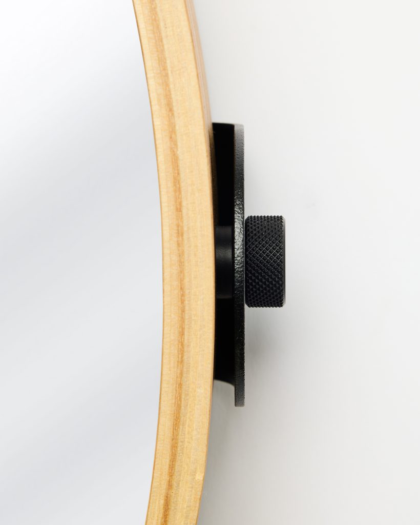 Close-up of a WoodUpp round mirror showing its pale layered wooden frame and the WoodUpp black powder-coated metal wall bracket with a textured knurled adjustment knob, revealing how the slim mirror sits offset from the wall on the compact mounting plate.