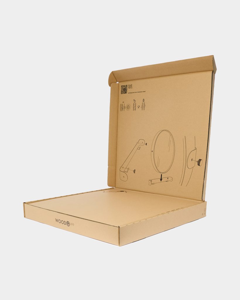 Open WoodUpp corrugated cardboard box photographed on a neutral background, WoodUpp logo printed on the front edge. The inside lid features line drawings and assembly instructions for a circular WoodUpp mirror, its wooden articulated wall arm/bracket, mounting plate and fixing screw.