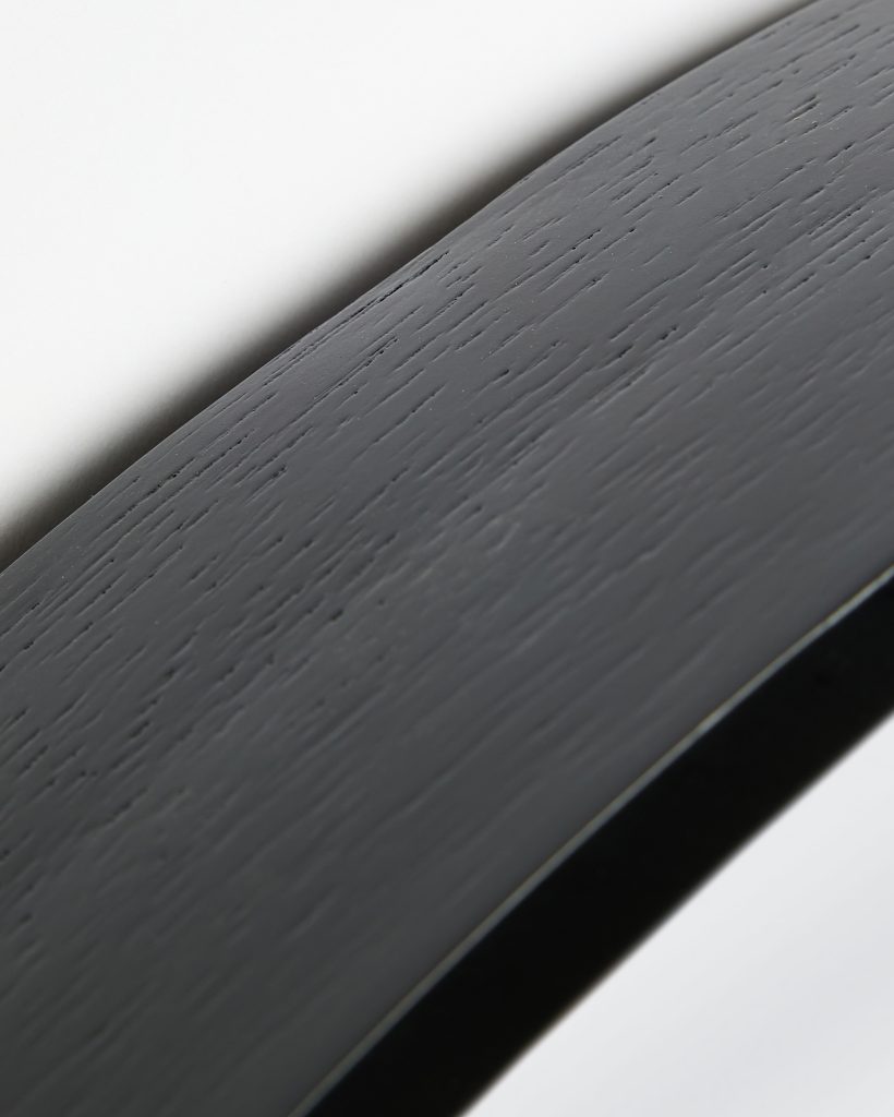 Close-up of a WoodUpp product: a curved, matte black wood surface showing pronounced oak grain texture. The photograph highlights the smooth painted finish and beveled edge of a WoodUpp wooden plank or veneer sample, emphasizing fine grain detail and subtle light reflection.