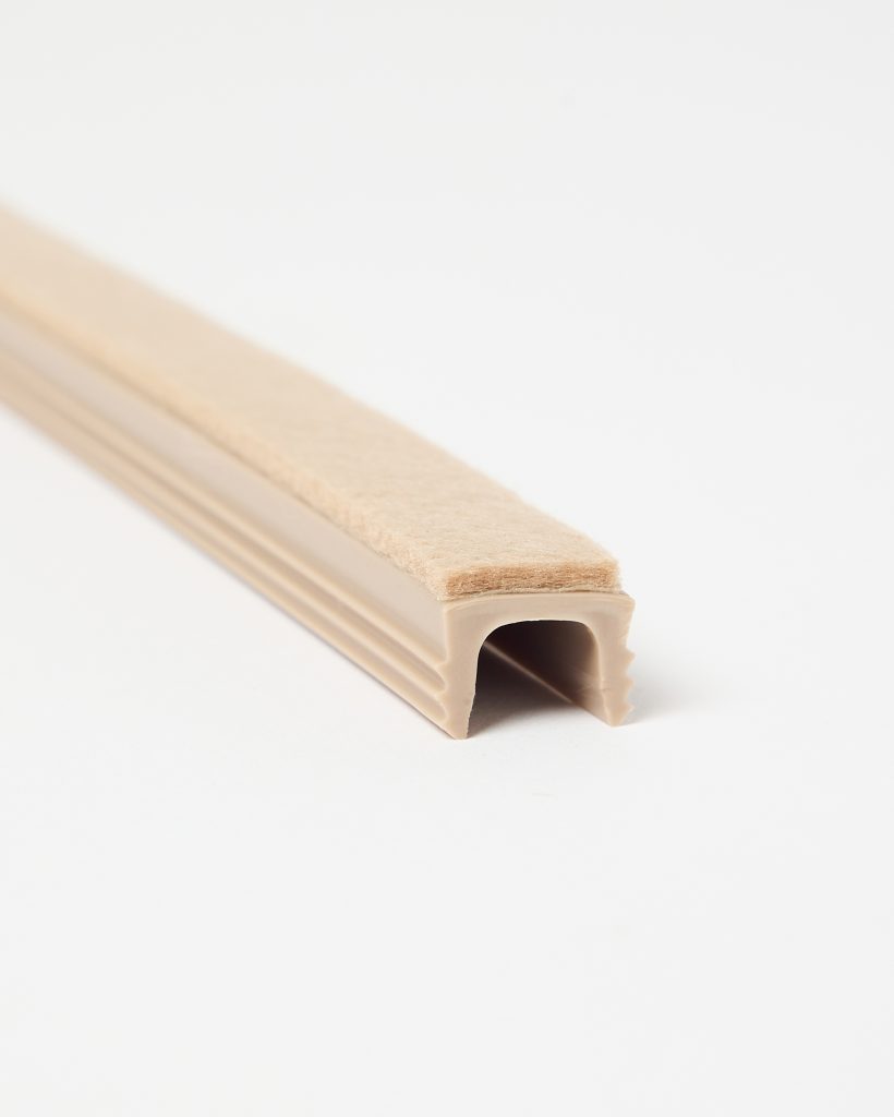 Close-up of a WoodUpp beige U-shaped plastic edge trim with an attached tan felt strip: a flexible channel profile designed to clip over doors or window edges, providing soft felt cushioning and draught-sealing. Textured ribs on the exterior and hollow interior are visible.