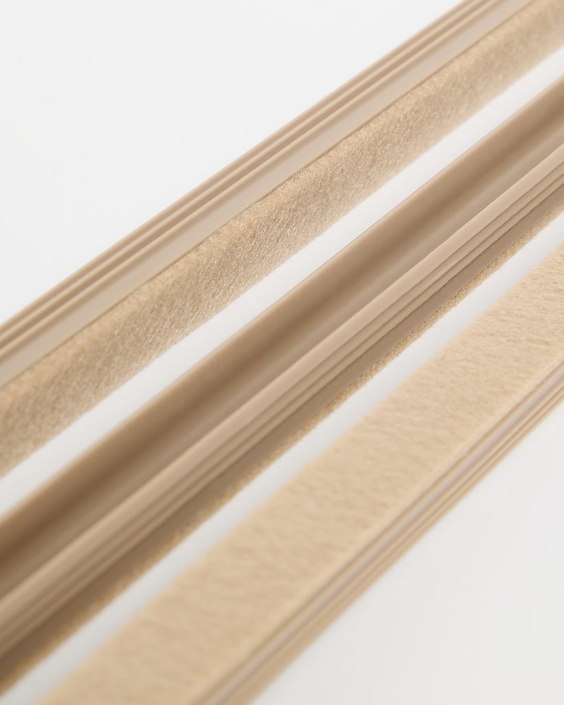 WoodUpp product photo showing three beige self-adhesive door seals: two felt adhesive strips and a central rounded rubber gasket with a ribbed PVC base. Close-up highlights soft felt pile, smooth flexible rubber and slim profile designed for draught-proofing gaps around doors and windows.