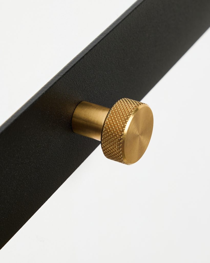 Close-up of a WoodUpp matte black metal panel fitted with a cylindrical, brushed brass knurled knob/coat peg. The gold-toned, textured circular head and short stem project from the plate, highlighting the knob’s fine diamond knurling and smooth machined finish and minimalistic contrast.