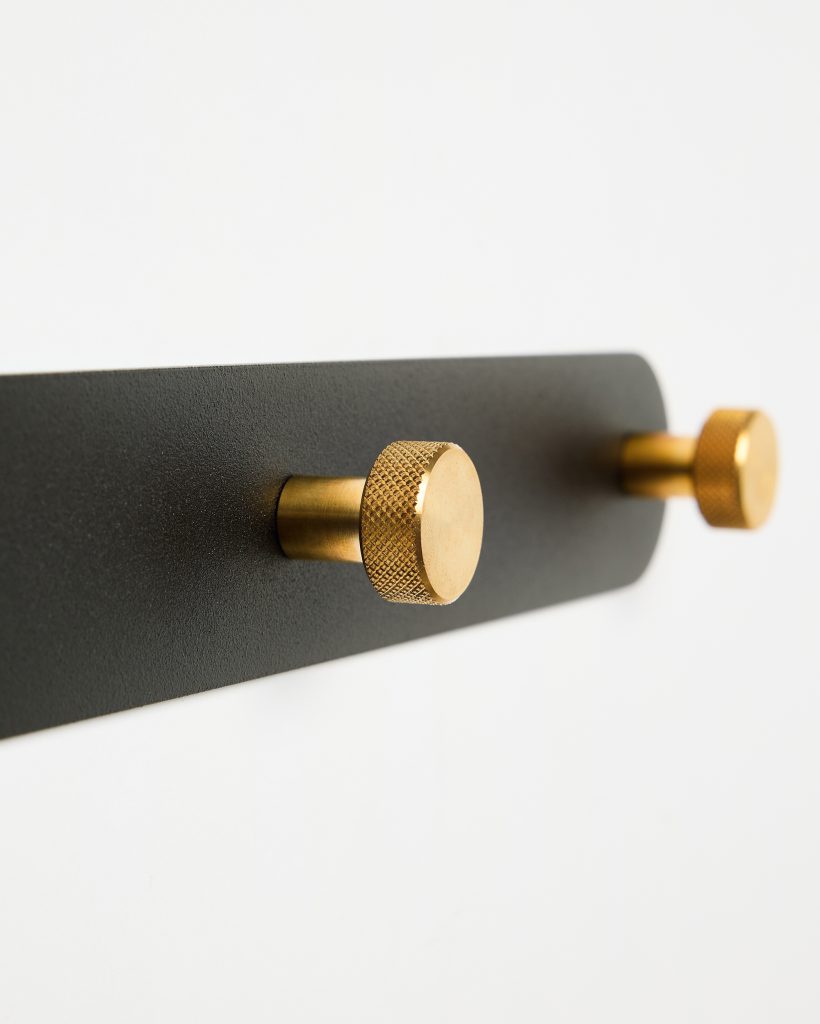 WoodUpp wall-mounted coat rack: matte black rectangular metal backplate with two gold-brass cylindrical pegs featuring knurled textured heads, short stems and polished finishes, shown close-up against a white wall—minimal, contemporary hook design suitable for hanging coats, bags or towels for everyday use.