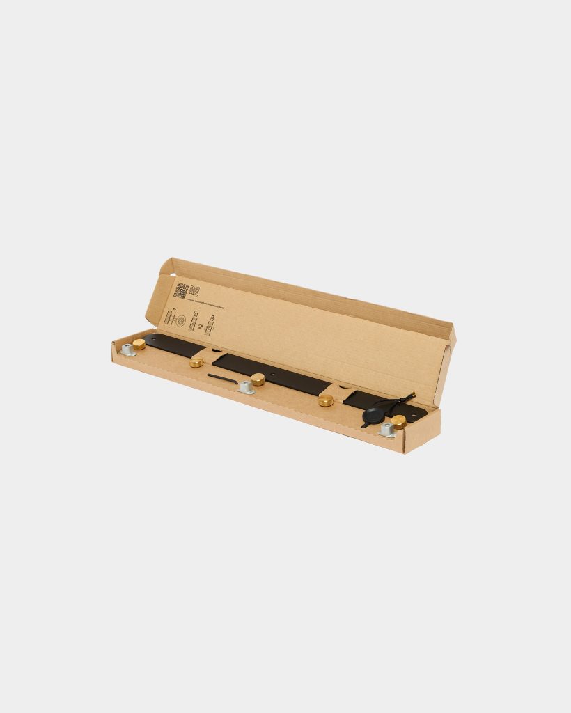 WoodUpp product: open cardboard box containing a black metal wall rail with three brass round knobs, two silver T-shaped mounting brackets and screws, plus a short black connector cable. Items are neatly fitted in recycled cardboard packaging, showing assembly hardware and finish details.