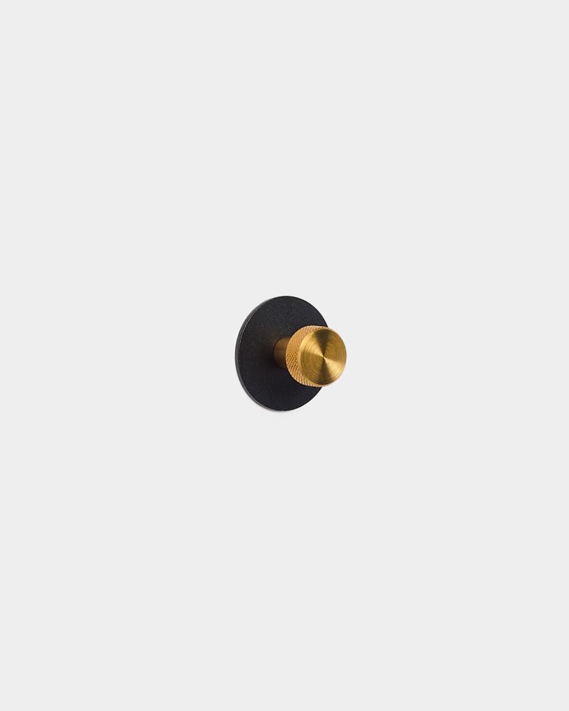 WoodUpp wall hook: a small matte black circular backplate mounted on a white wall, fitted with a cylindrical brass-gold knob featuring a knurled textured face and brushed finish. Minimal, modern coat hook/peg design from WoodUpp, shown centered against a clean light-gray background.
