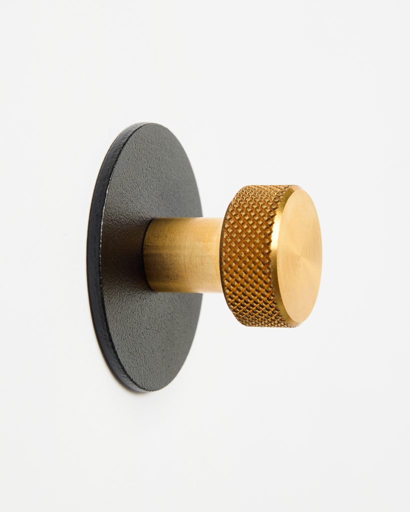 Close-up of a WoodUpp wall hook: a textured, knurled solid brass knob and short brass stem protruding from a matte black round base plate mounted on a white wall. The hook’s gold-toned finish and industrial knurled grip contrast with the flat black disc backing.