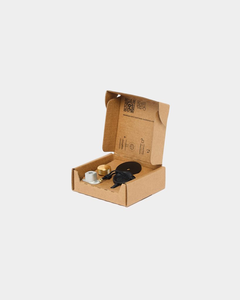 Open WoodUpp cardboard box displaying printed installation icons and a QR code, containing WoodUpp hardware: a brass knurled metal knob, a silver hexagonal mounting insert, a black circular rubber gasket/plate, and a short black braided cord with a small black leather tag — neatly arranged inside.
