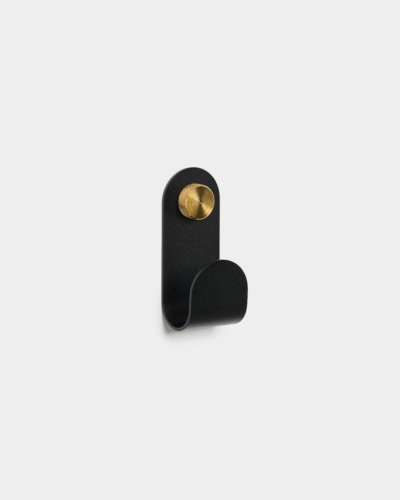 WoodUpp wall hook: a minimalist black powder-coated metal J-hook mounted on the wall, featuring a curved loop for hanging and a textured brass knurled mounting screw. Clean Scandinavian design with matte black finish and warm brass accent, shown isolated on a light grey background.