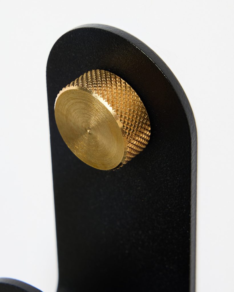 Close-up of a WoodUpp matte black metal wall hook featuring a rounded top and a circular brass knurled knob; the textured golden knob protrudes as a decorative peg for hanging, contrasting the smooth black finish and highlighting precise machining and minimalist modern hardware design.