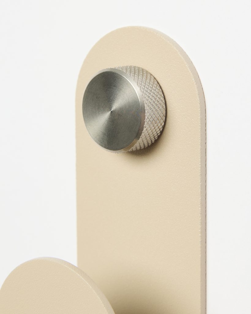 WoodUpp close-up of a beige wall hanger: a rounded rectangular panel and partial circular hook finished in matte cream, topped by a textured stainless-steel knob with knurled edge and flat brushed face. The minimalist WoodUpp wall hook emphasizes clean lines and tactile metal detailing.
