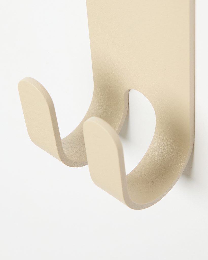 Close-up of a WoodUpp beige metal double wall hook: a rectangular backplate with two U-shaped prongs ending in flat tips, featuring a smooth matte finish and gentle curves, mounted on a white wall. Designed for hanging coats, bags or towels.