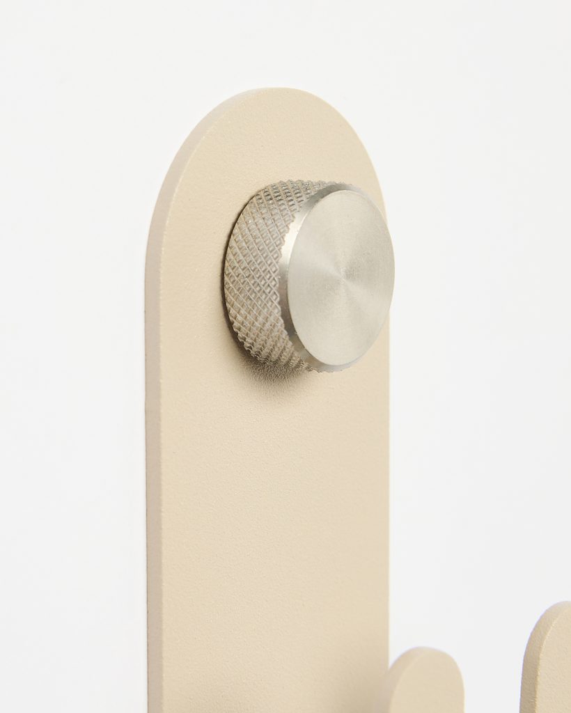 Close-up of a WoodUpp wall hook: a matte beige vertical backplate with rounded top and a brushed, knurled stainless-steel knob mounted as the hanging peg. The image shows textured paint, the metallic screw-like cap, and a partial second beige hook against a white background.