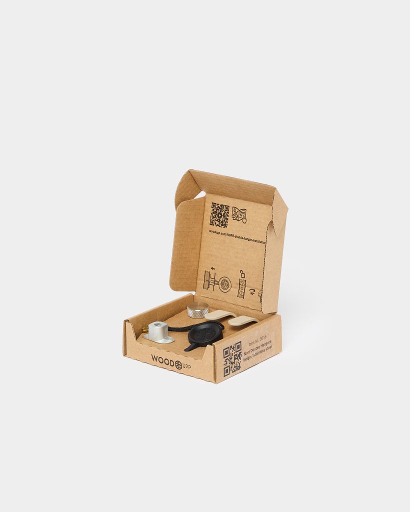 WoodUpp open cardboard box displaying mounting hardware: a knurled silver metal knob, a small metal mounting stud, a black circular magnetic disc on a braided cord, two beige adhesive wall clips and assorted screws — all neatly arranged inside branded eco cardboard packaging.