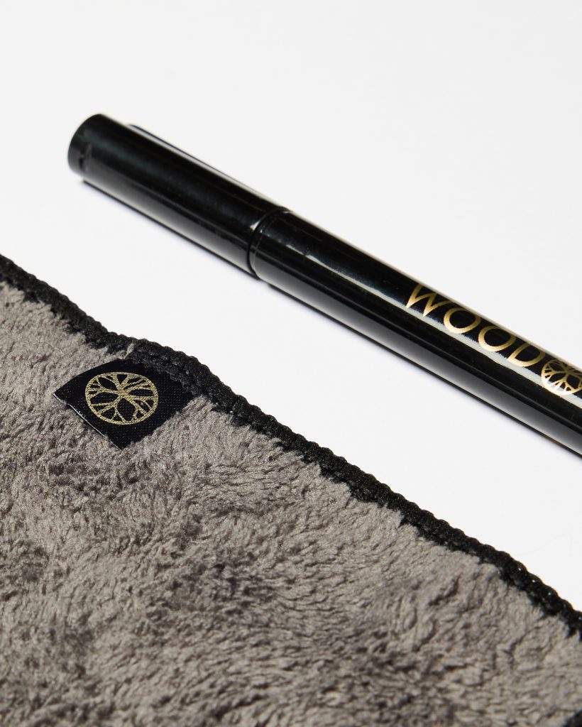 Close-up on WoodUpp products: a glossy black pen-like applicator printed with gold "WOODUPP" branding and a soft plush grey microfiber cloth with black stitched edge and a small black tag bearing WoodUpp's gold circular logo, photographed against a white background.