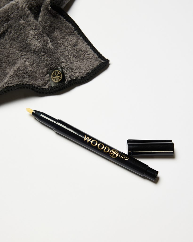 WoodUpp black applicator pen with gold "WOODUPP" logo and beige sponge tip lies uncapped beside its black cap, accompanied by a grey plush microfibre cloth with black trim and a small circular gold WoodUpp logo tag, photographed on a white background.