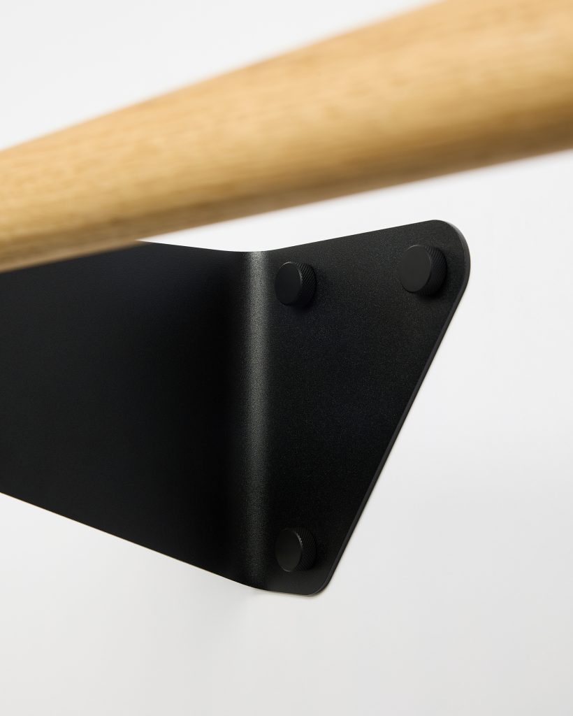 Close-up of WoodUpp hardware showing a light oak handrail mounted on a matte black triangular wall bracket. The WoodUpp bracket features three circular knurled mounting caps and a folded-steel profile, contrasted against a white wall, highlighting the join between the wooden rail and minimalist metal fitting.