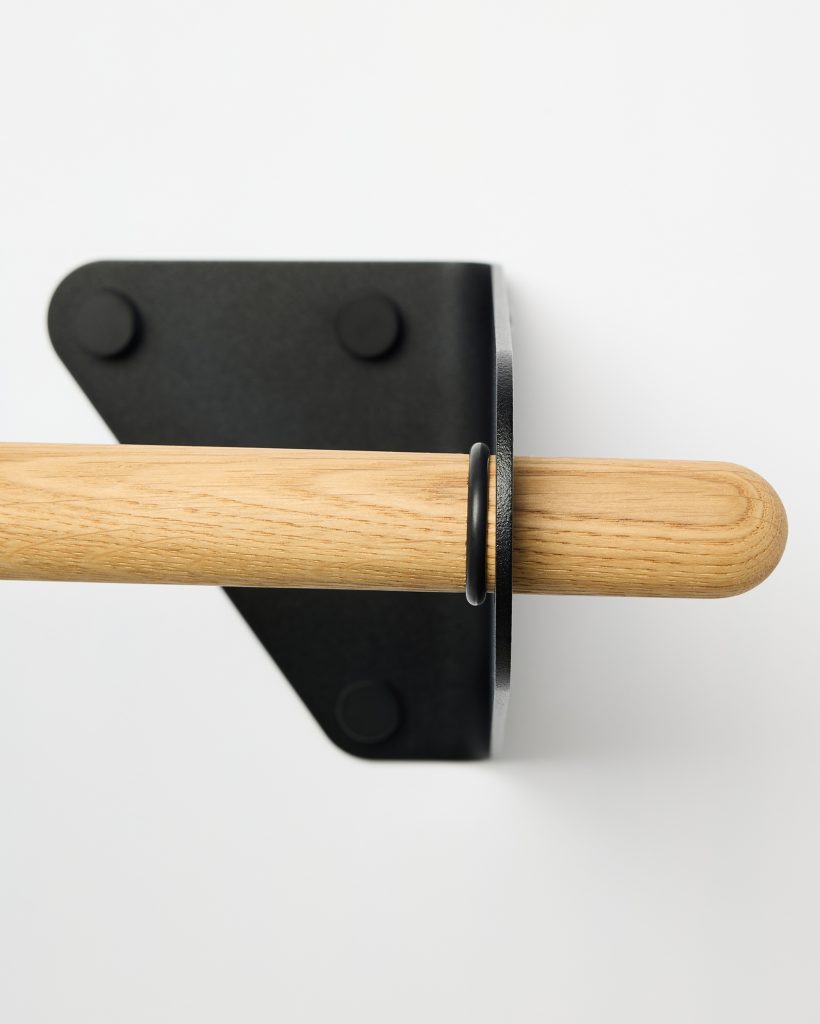 Close-up of a WoodUpp oak rail and matte black steel wall bracket: a smooth, rounded oak dowel showing natural wood grain and satin finish, held by a minimal black triangular bracket with rubber pads and a slim black metal collar, mounted against a white wall.