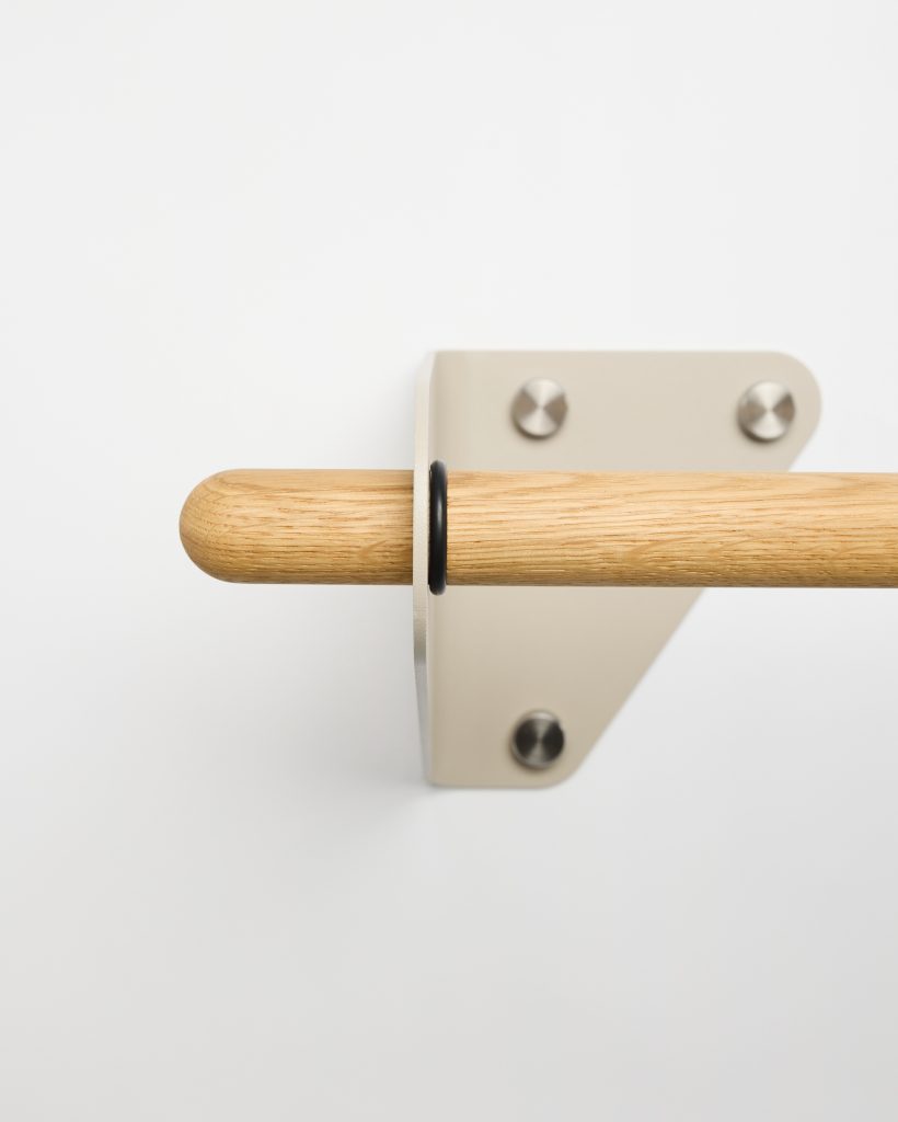 WoodUpp oak handrail with rounded end mounted on a beige metal wall bracket is shown; stainless screws fix the triangular bracket, and a black rubber spacer ring secures the rail. Clean white background highlights the minimalist WoodUpp wooden rail and matching hardware.