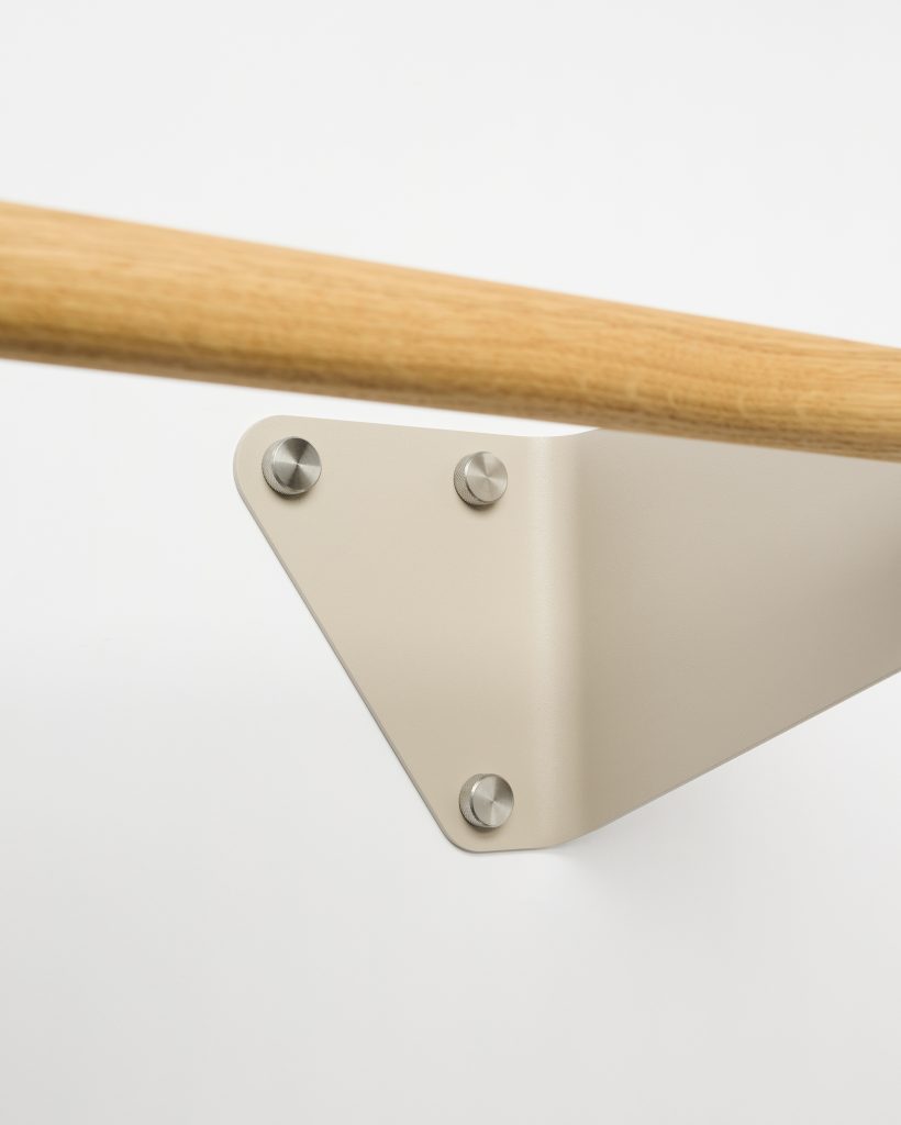 WoodUpp wall-mounted handrail: close-up showing a smooth oak wooden handrail fastened to a pale beige triangular metal wall bracket by three round stainless-steel bolts. The minimalist bracket’s folded steel plate and oak rail highlight WoodUpp’s clean, Scandinavian-inspired hardware and timber finish.
