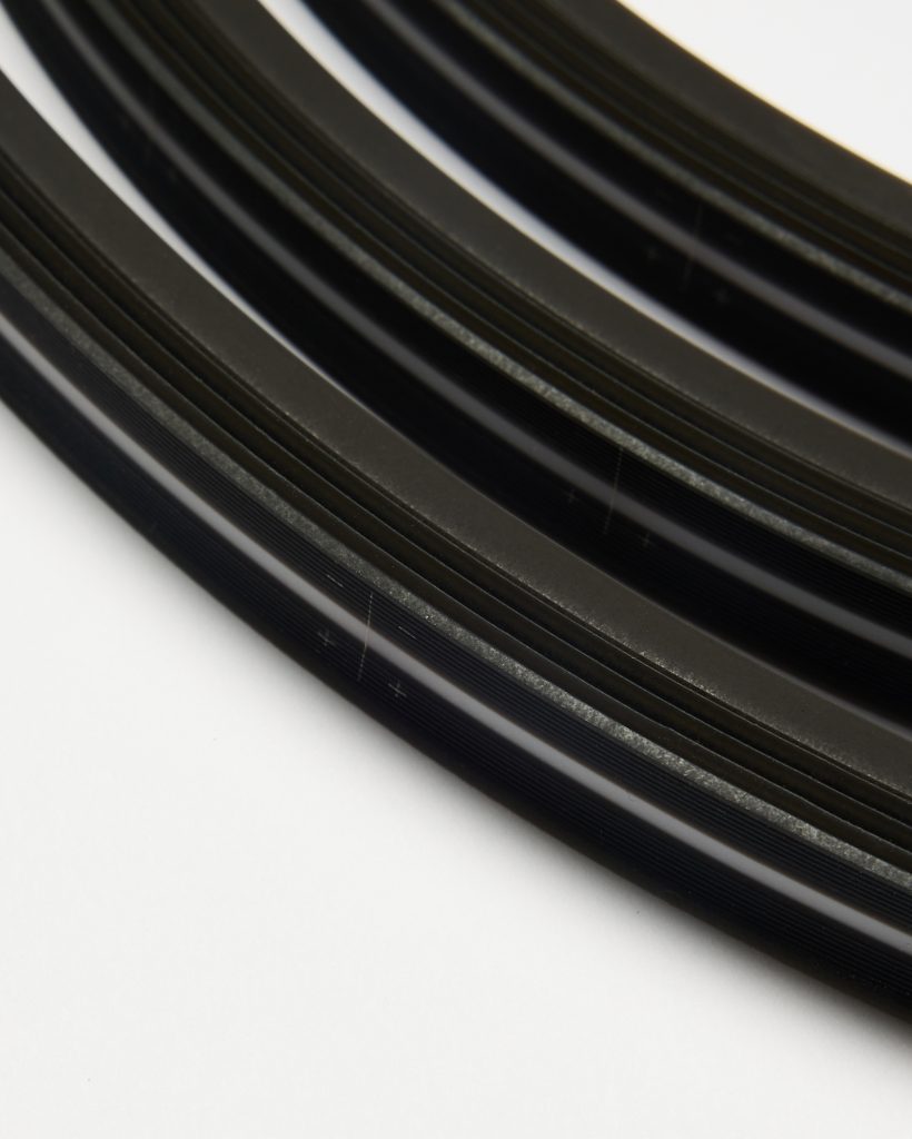 Close-up of WoodUpp sanding belts: several black abrasive belts with fine longitudinal ribs and a thin white alignment stripe, coiled and stacked in parallel arcs on a white background, showing textured grit surfaces and smooth backing edges suitable for belt sanders.