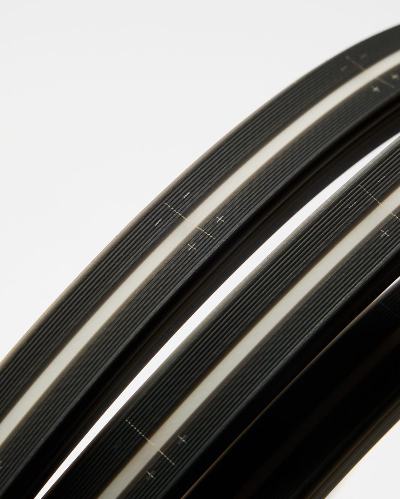 Close-up of WoodUpp black rim tapes — three curved, ribbed rubber strips with a central light-grey reflective stripe and tiny printed alignment crosses, shown against a white background. The WoodUpp rim tapes appear textured for grip and protection, with smooth, rounded edges.