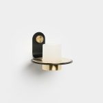 WoodUpp wall sconce featuring a matte black wall bracket with a knurled brass mounting knob, a flat black-and-brass circular shelf, and a frosted cylindrical glass shade set over a brass cup — an elegant minimalist, mixed-metal wall lamp from WoodUpp.