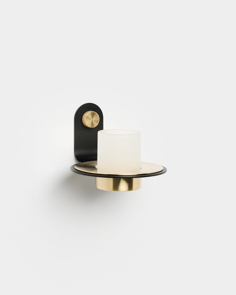 WoodUpp wall sconce featuring a matte black wall bracket with a knurled brass mounting knob, a flat black-and-brass circular shelf, and a frosted cylindrical glass shade set over a brass cup — an elegant minimalist, mixed-metal wall lamp from WoodUpp.