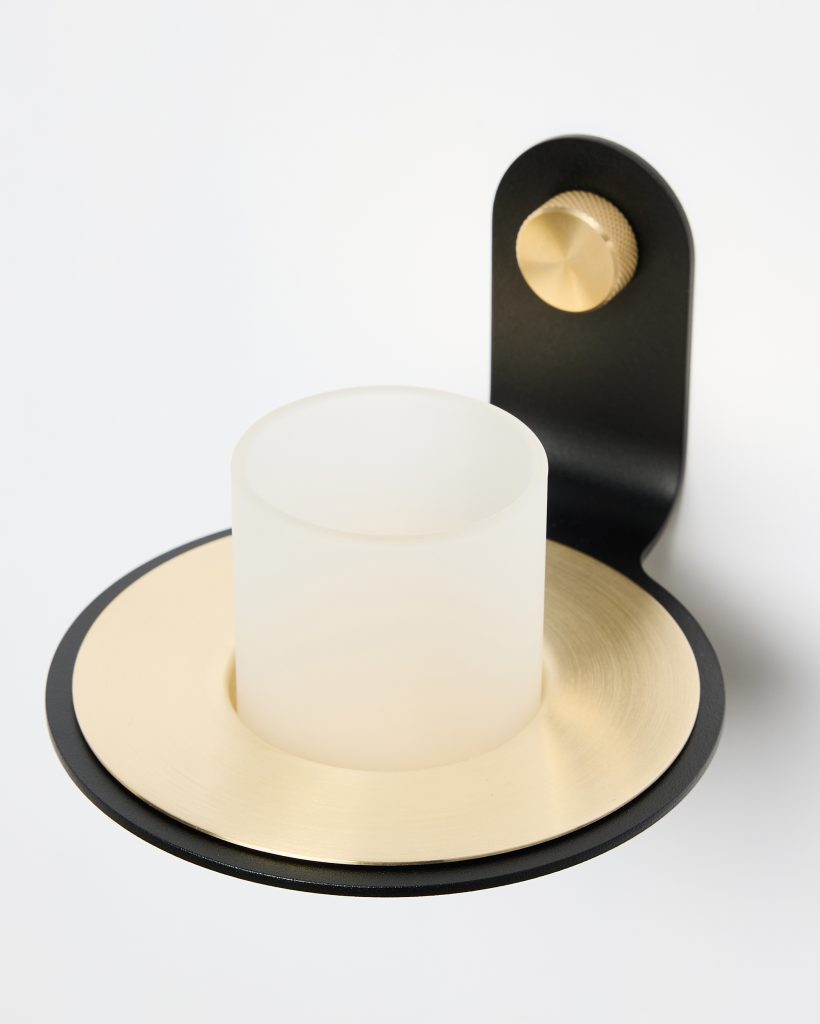 WoodUpp wall-mounted candle holder: a matte black metal bracket with knurled brass adjustment knob supporting a circular brushed brass plate and a frosted cylindrical glass votive cup, shown close-up against a white background, highlighting the mixed brass and black finish and minimalist modern design.