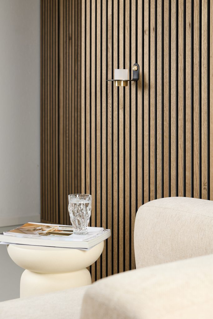 WoodUpp oak slat wall panels form a vertical, evenly spaced natural wood feature wall. A small black-and-brass WoodUpp wall candle holder with a white votive is mounted on the slats. In front, a cream sculptural side table holds magazines and a textured glass beside a beige upholstered sofa.