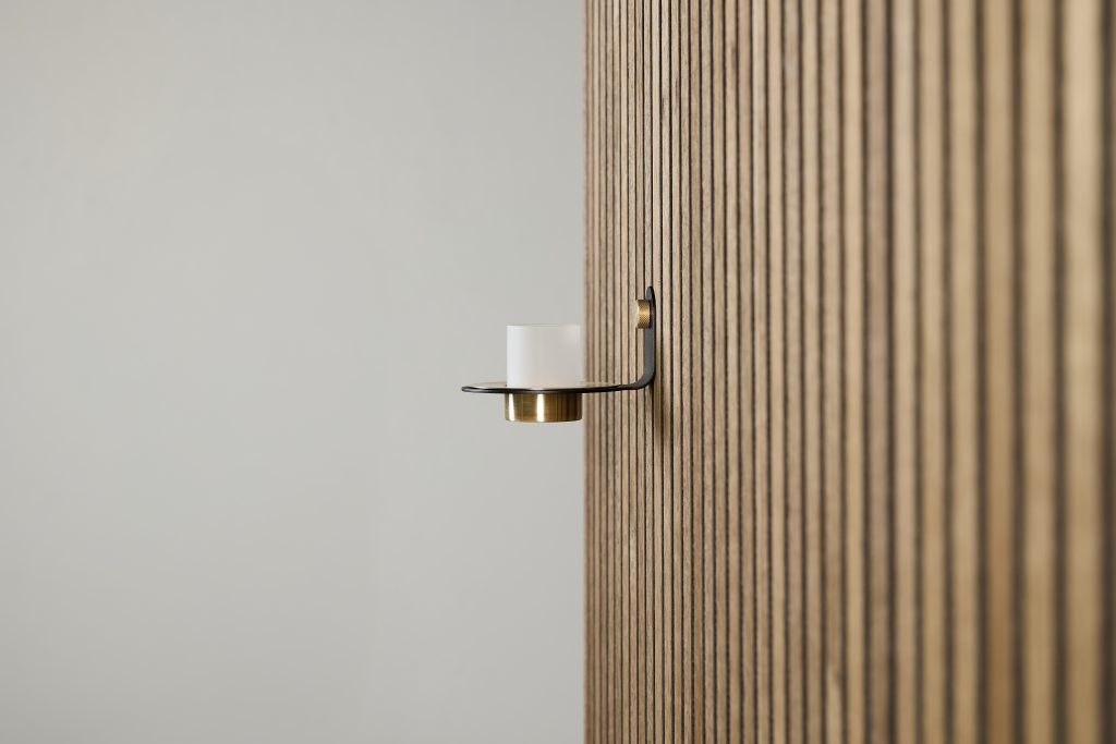 WoodUpp wall display showing a minimalist WoodUpp wall-mounted candle holder: a black metal arm with brass cup holding a frosted glass votive, attached to a vertical oak slatted WoodUpp panel. Neutral grey background highlights the clean lines and warm wood-grain texture.