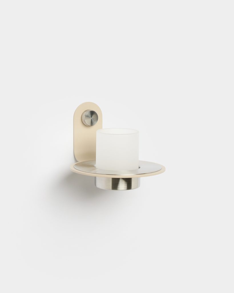 WoodUpp wall sconce: a beige wall-mounted bracket with knurled stainless knob supporting a circular brushed-metal tray and recessed holder, topped by a frosted cylindrical glass votive. It features an elegant matte finish and compact profile. Unlit, mounted on a white wall, showcasing modern Scandinavian design.
