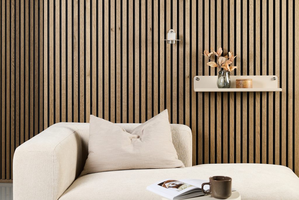 Minimal living area with WoodUpp oak slatted wall panels in vertical dark-groove pattern, paired with a beige WoodUpp floating wall shelf mounted with visible metal fixings holding a glass vase and wooden jar; cream upholstered sofa, neutral cushion, open book and coffee cup on the chaise.